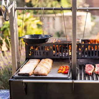 Outdoor grill with food being cooked, including bread and vegetables, in a garden setting.